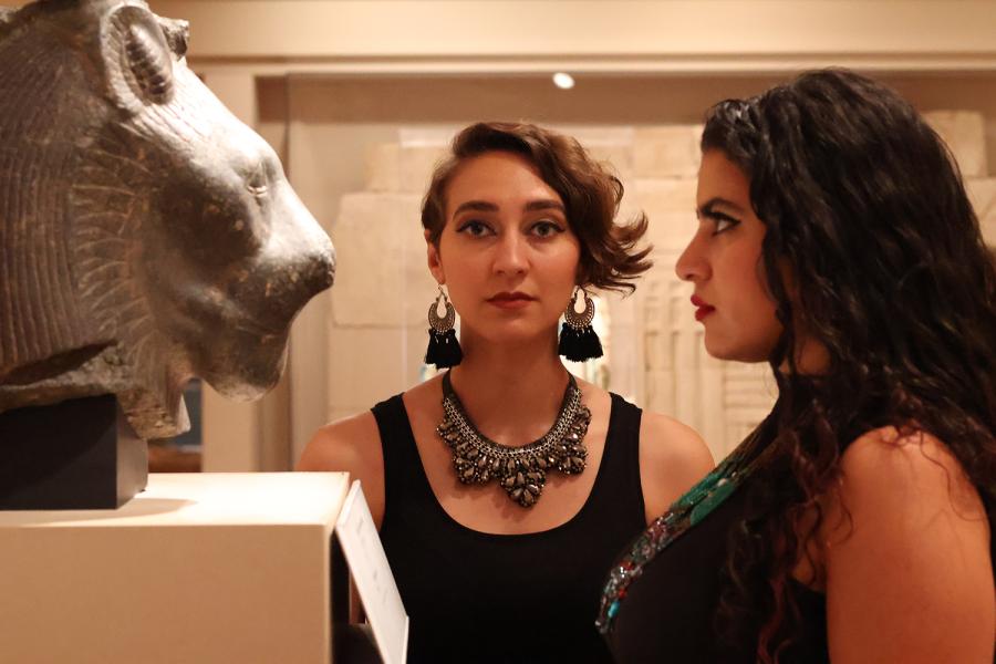 In a gallery, two women pose with a lion statue. One faces the lion head on while the other looks into the lens.