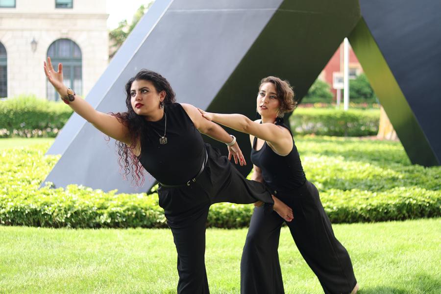 On a museum lawn, two women dance. One leans forward while the other leans on her back.