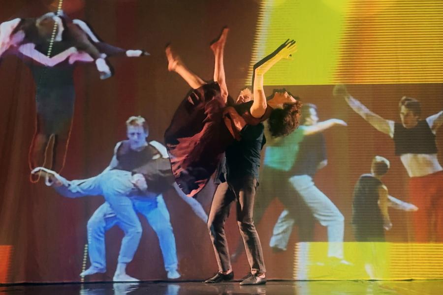 In front of a background covered in projections of dancers, one performer picks up another.