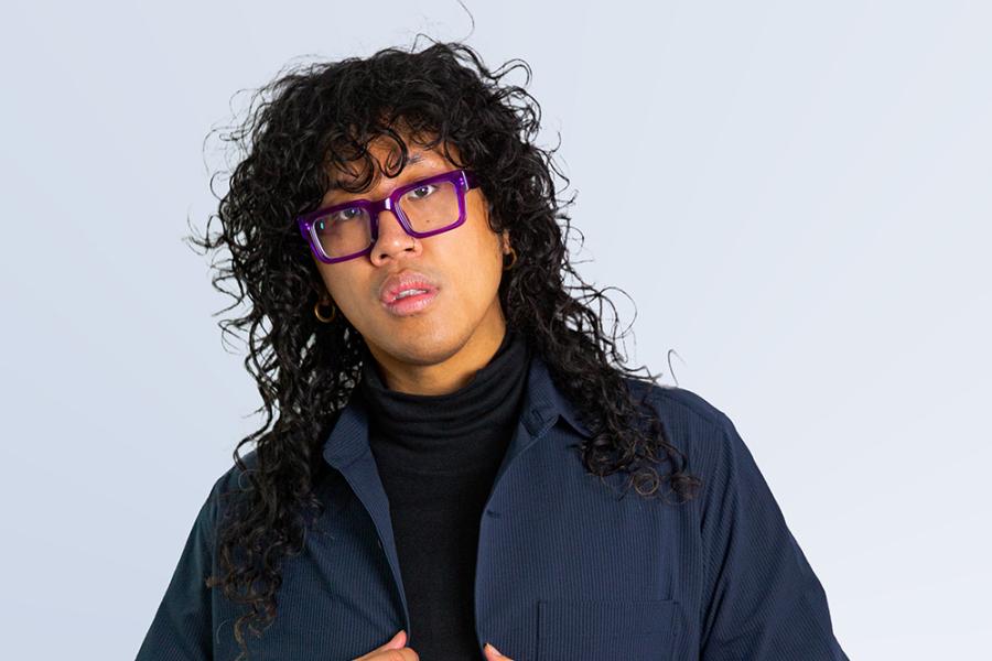 A person with long black hair and thick-framed glasses holds their navy jacket in front of a white backdrop.