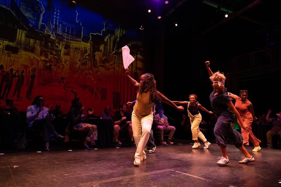 On a stage, four women tap dancers, in plain clothes, perform with their arms out.