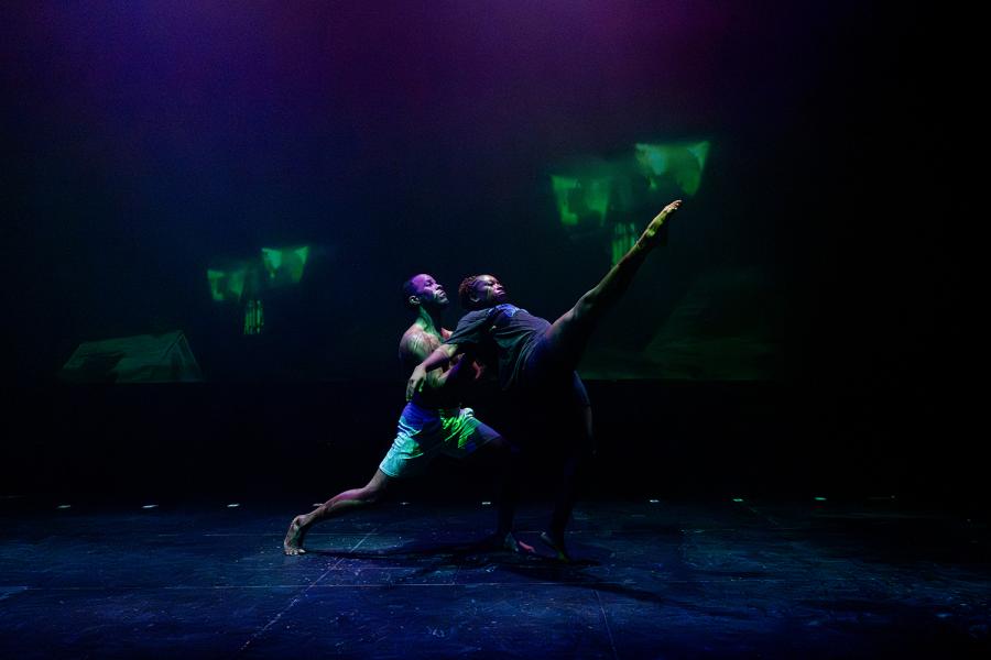 In dark lighting, one female dancer throws her leg up and leans back on a male dancer.