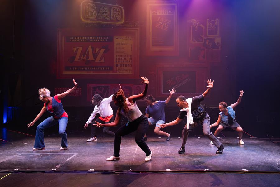 Seven tap dancers on a stage. They lean to the left with their right hands up.
