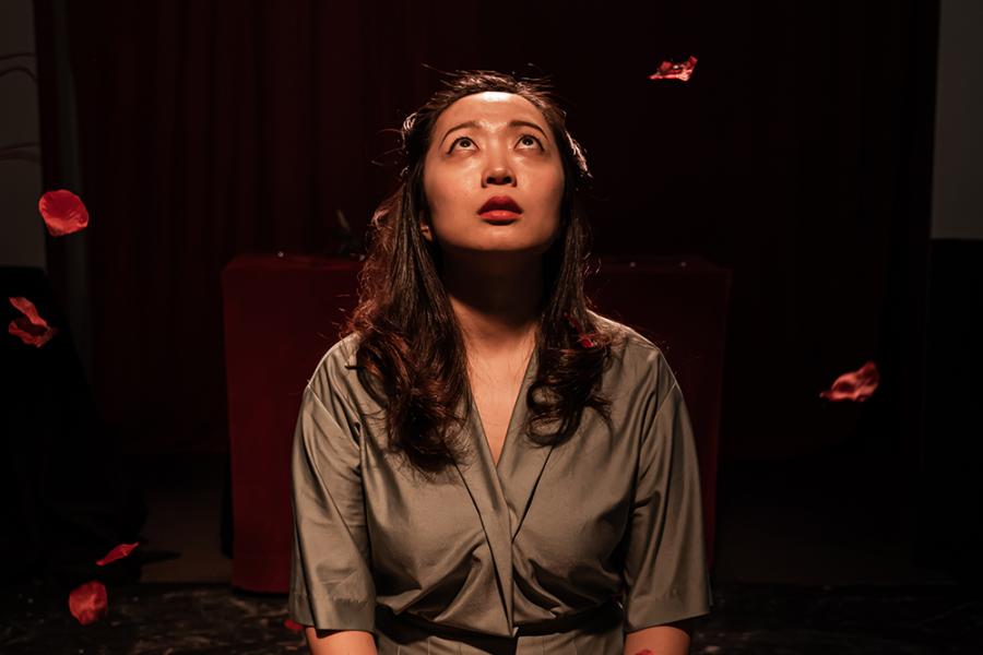 A young Japanese women kneels and prays in a spotlight. She has long brown hair and wears a grey/green wrap dress. Red rose petals fall around her.