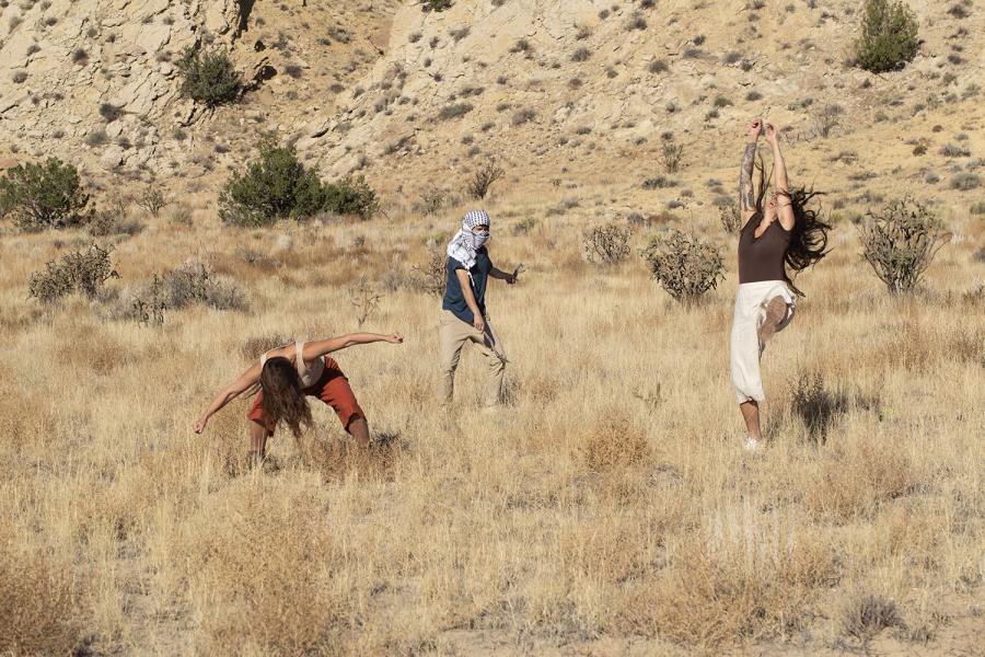 In the desert, three women dance in casual attire. One has a keffiyah wrapped around her head.