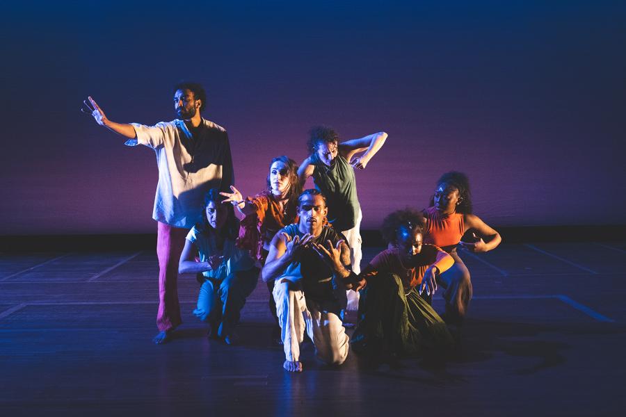 On a stage with dramatic lighting, a group of dancers face the audience with their heads in an almost diamond formation. One outlier stands to the left above them.