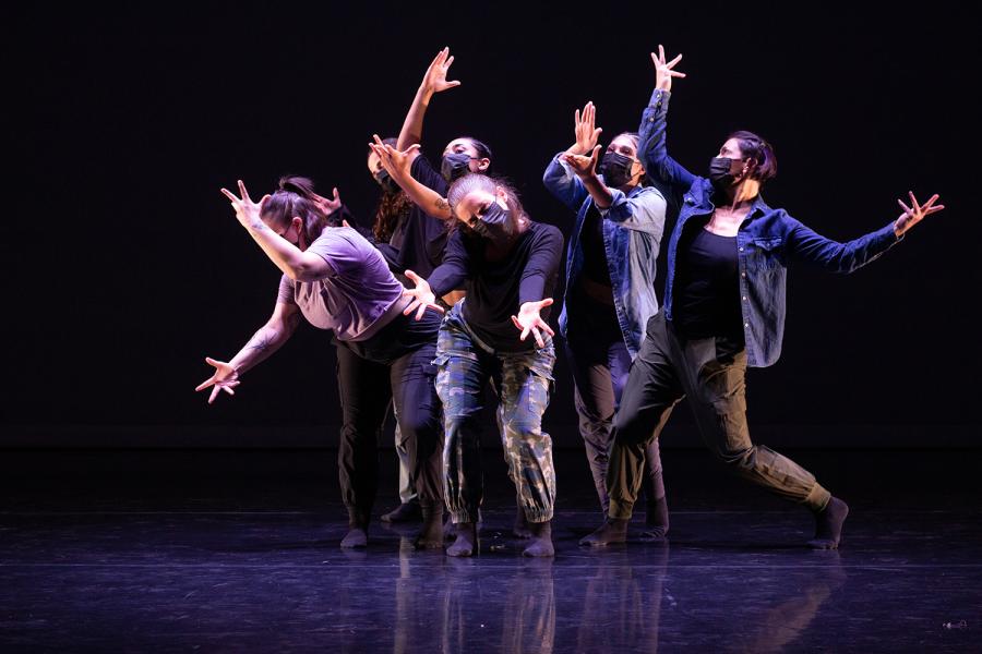 On a stage, with spotlights, five dancers stand close to each other and throw their arms out wide.