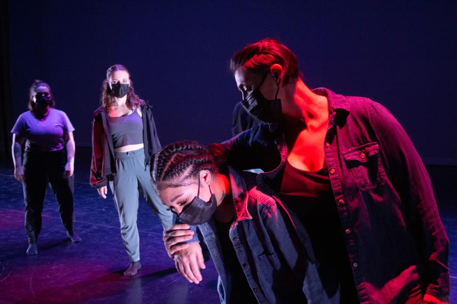 In the foreground, one dancer has their arm around another dancer. Two dancers watch in the background. They all wear face masks.