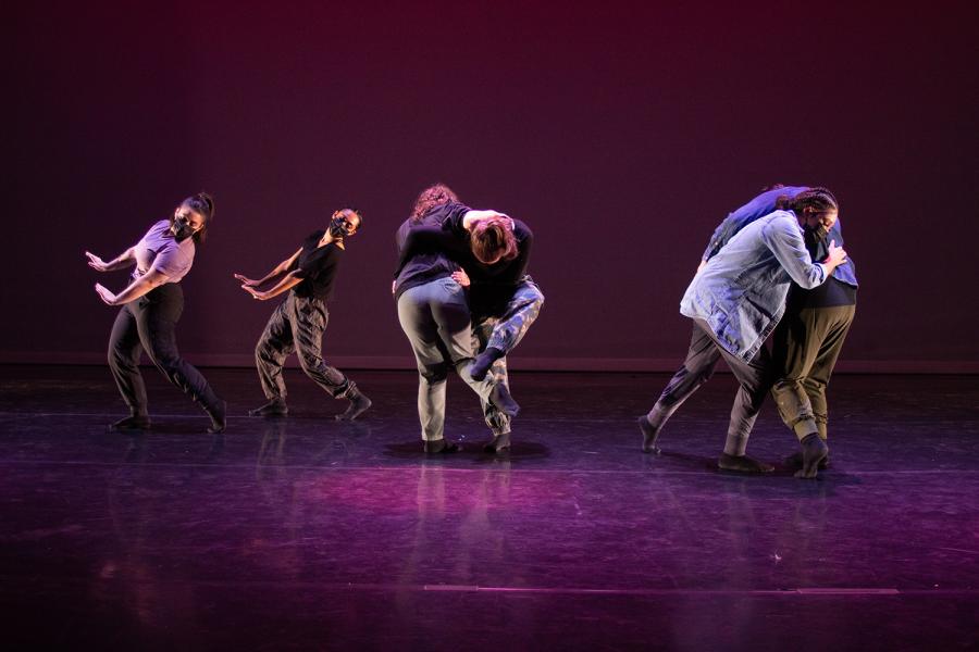 Six dancers on a stage with purple spotlights. Two huddle. One lifts another and two gesture with their hands like they're exiting stage right.