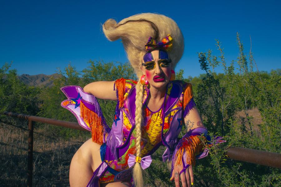 A drag queen in a colorful purple vest leans on a railing. There is green foliage and a clear blue sky in the background.