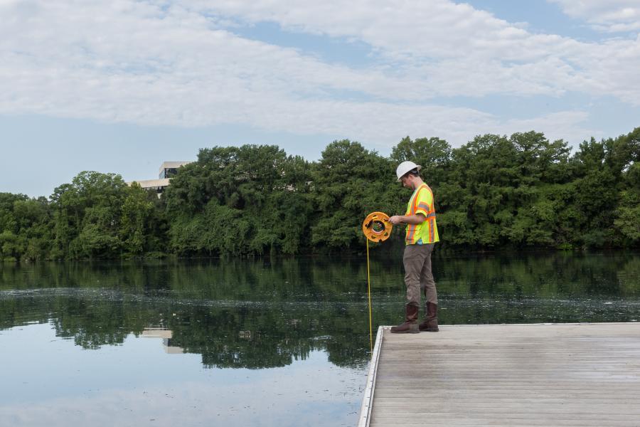 By a body of water, a man, in a hard hat and orange vest, fishes.