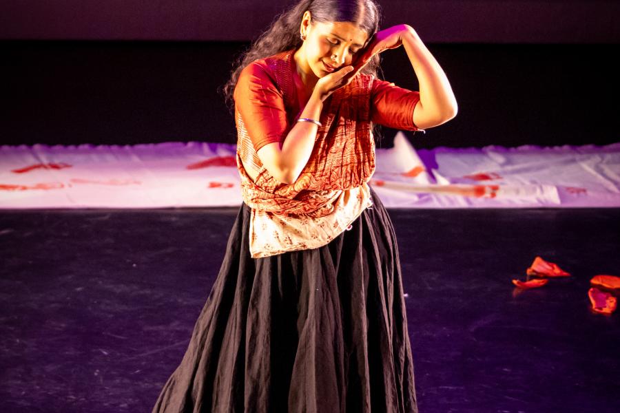 A performer in a long dark skirt holds the side of their face with their hands.