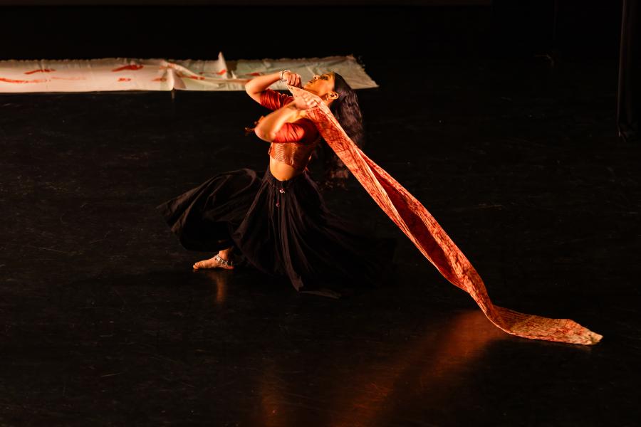A performer in a dark skirt drags a piece of orange cloth across a stage.