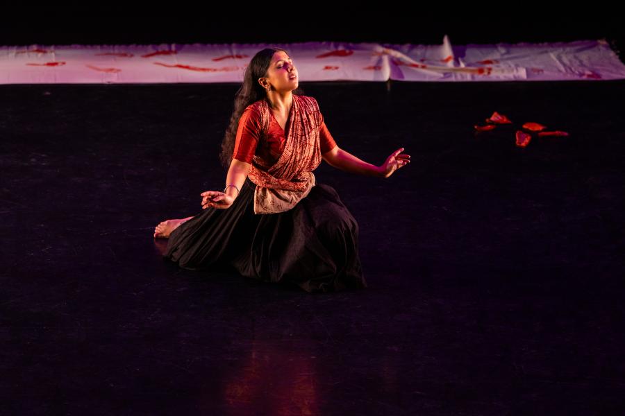 A performer in a long dark skirt kneels on a stage with their eyes closed.