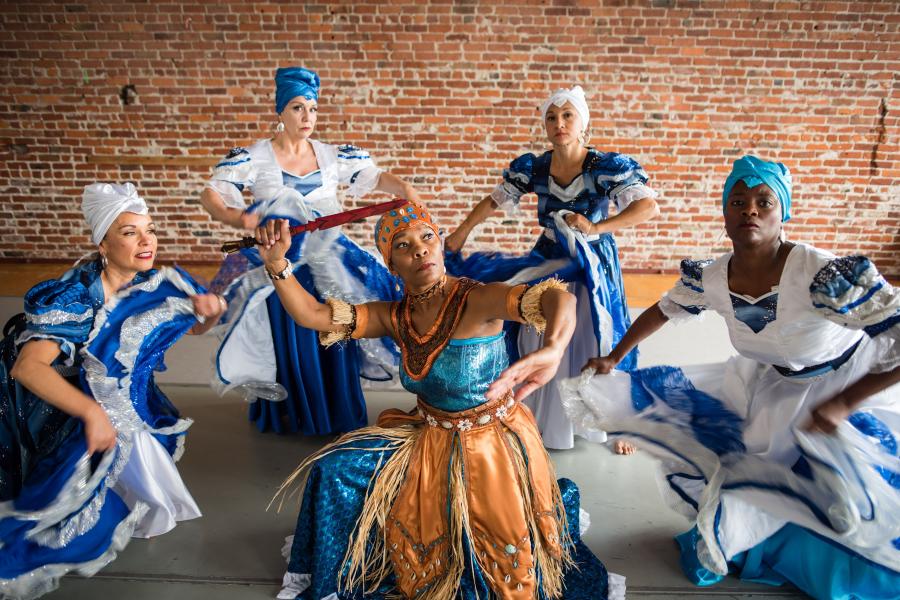 Five Afro-Cuban women dance in white and blue dresses and white and teal head wraps.