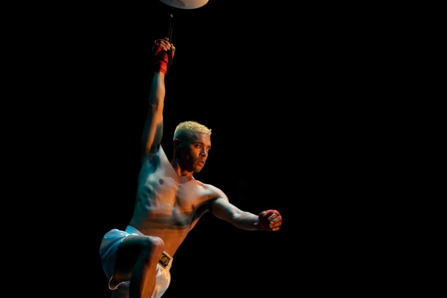 A shirtless, brown man, with blonde hair, swings from a punching bag in a spotlight.