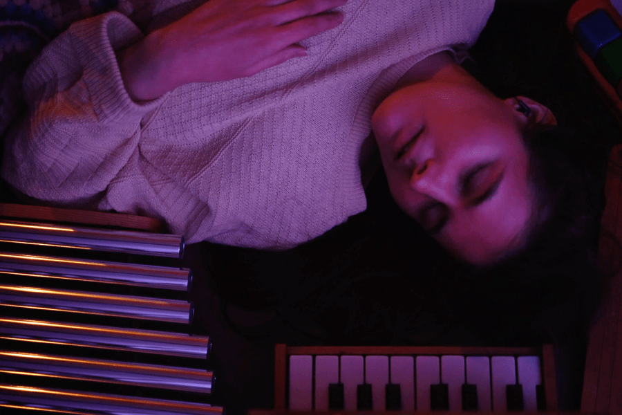 alexa dexa, a queer, non-binary, Sick, Mad, and Disabled white person, lays in bed in a textured yellow sweater, cozy under a crocheted blanket their γιαγιά made. Encircling them, from left to right, are their toy chimes, Michelsonne toy piano, toy zither, and tap-a-tune xylo-keyboard. alexa is restful, dreaming. Their left hand rests on their chest. Their long brown hair floats around them, indistinguishable from shadow. The light meeting their face is magenta, with light blue highlights.
