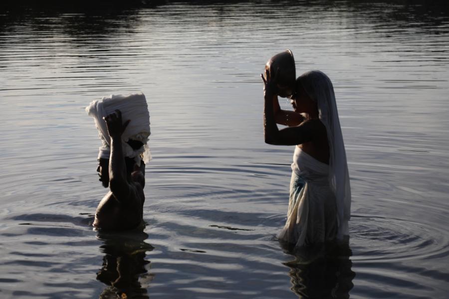 Two Black women wade in water. One holds a rock to her temple and the other has a white basket on her head.