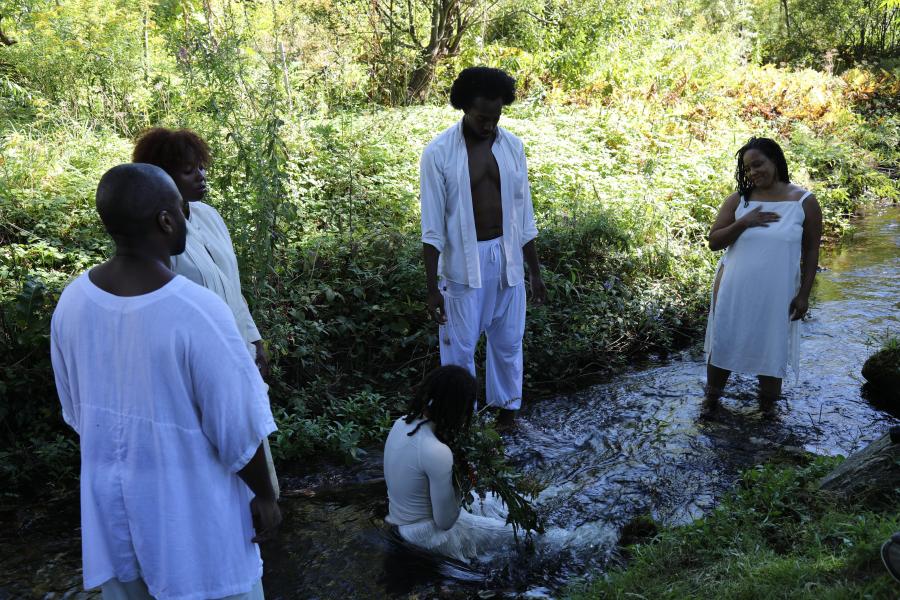 Four Black folks stand around another Black person who sits in a rocky river. They all wear white.