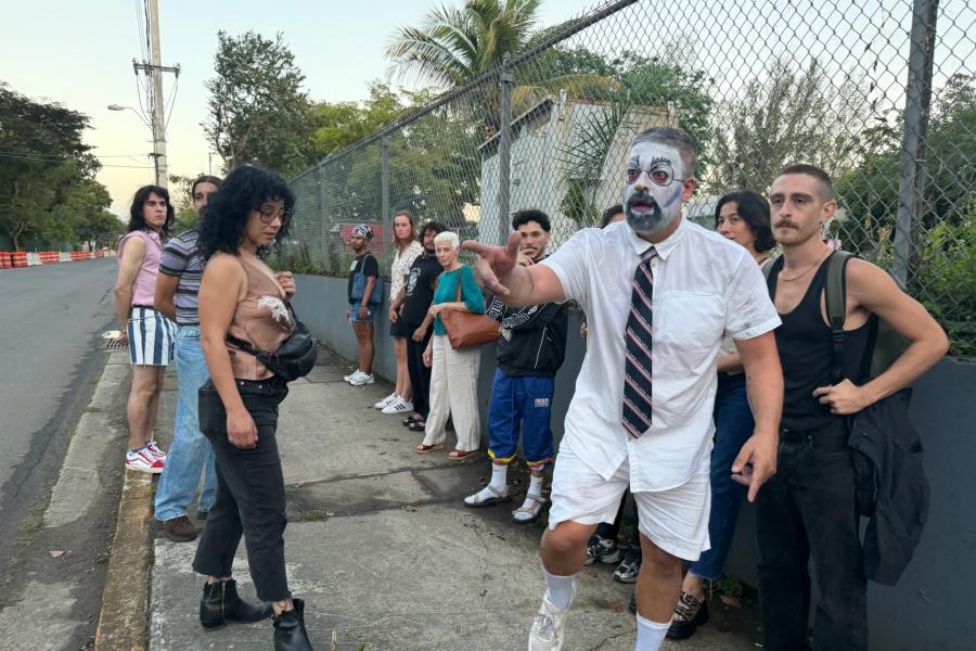 A person whose face has been painted white stands on a street, pointing at something offscreen. Eleven people look in the direction he's pointing.