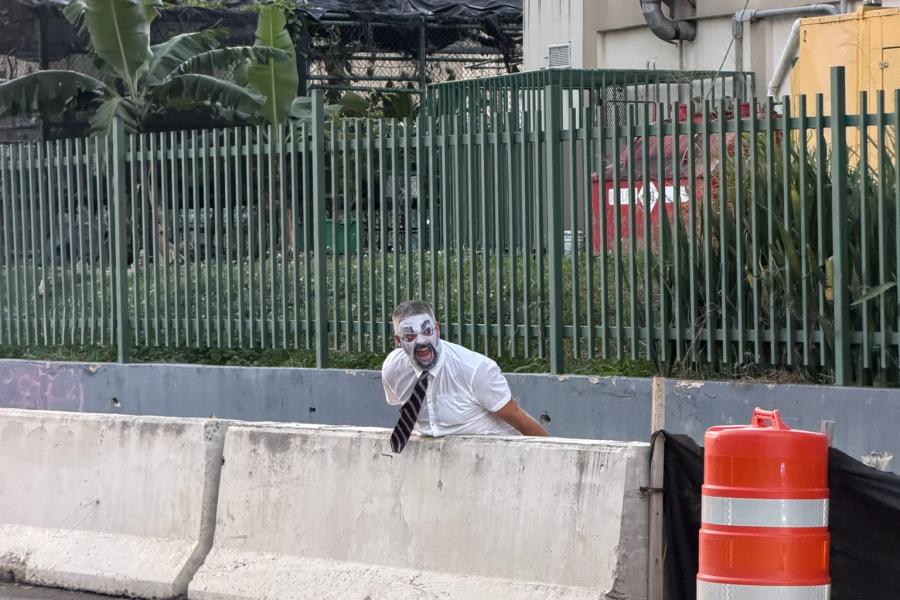 A person wearing a white shirt and tie whose face has been painted white is yelling by the side of a road.