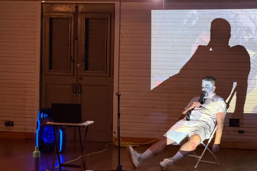 A person whose face has been painted white is sprawled out limply on a chair while holding a microphone to their mouth.