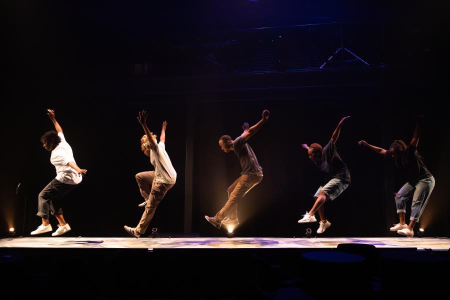 Five performers on a stage are mid leap with their arms raised over their heads and extended outwards.
