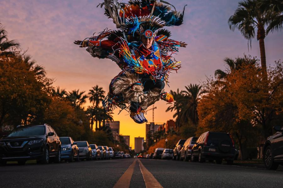 On a street, a dancer, in traditional Indigenous performance garb, jumps.