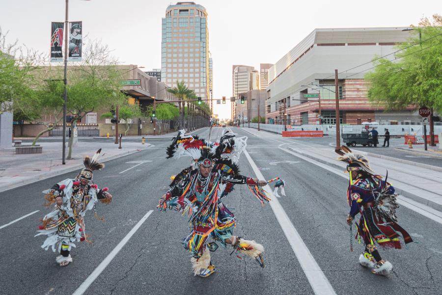 On a city street, three dancers, in traditional Indigenous garb, lean and hold their arms out in motion.