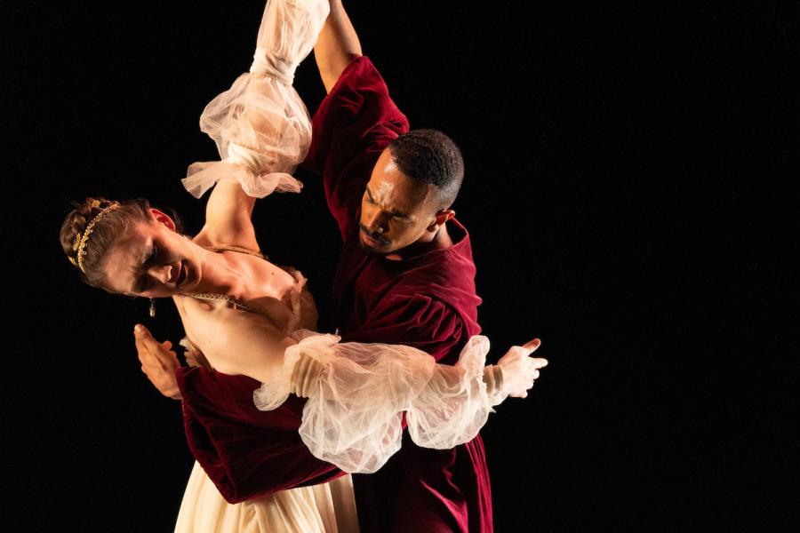 Two people in anachronistic clothing do a waltz in front of a black backdrop.