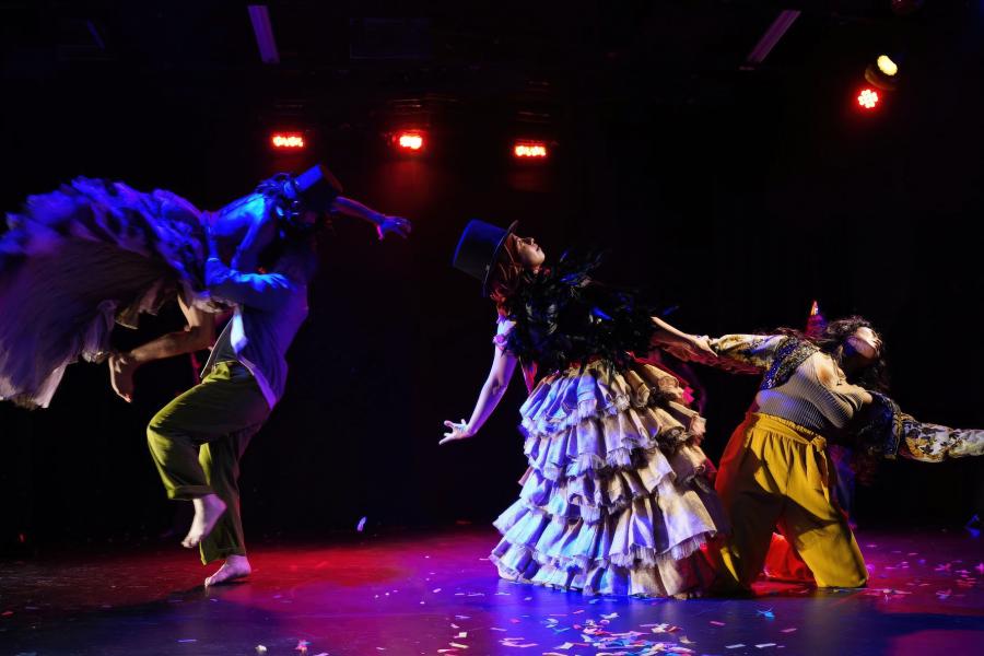 Two figures in frilled dressed dance with partners on a stage.