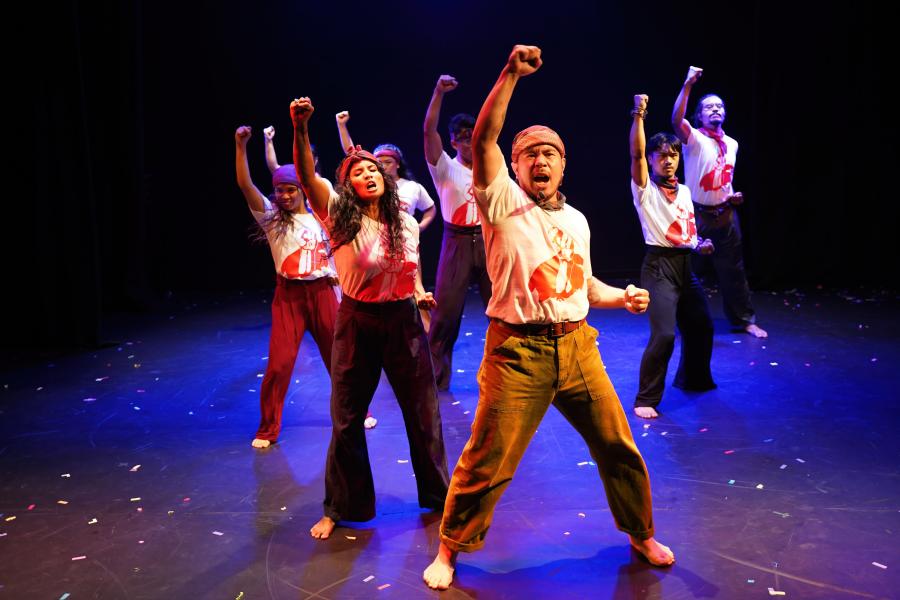Seven performers wear white shirts with red fists on them. They raise fists towards the sky.