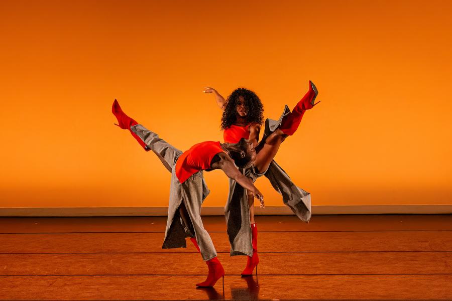 On a stage, with an orange backdrop, two dancers, in orange tank tops and grey skirts, spread their arms and legs. One lay throws their head back and leg out away from the other dancer.