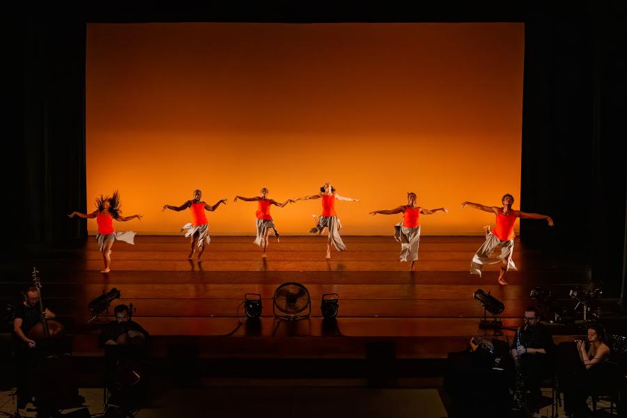 On a stage, with an orange backdrop, six dancers, in orange tank tops and grey skirts, spread their arms in a line. In the pit of the stage and foreground of the image, a live band performs.