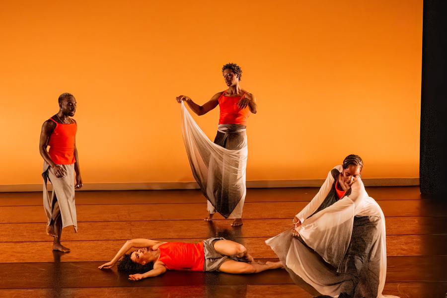 On a stage, with an orange backdrop, four dancers, in orange tank tops and grey skirts, perform. One lays flat on the stage.