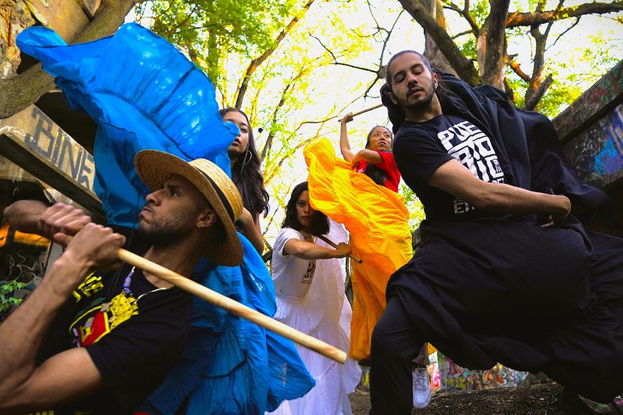 In the foreground, two men wield sticks, while three women in flowy gowns dance behind them. They're under trees.