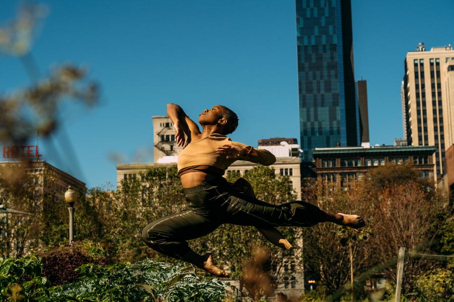 A Black dancer leaps on a city roof. Ivy covers buildings behind them on a sunny day.