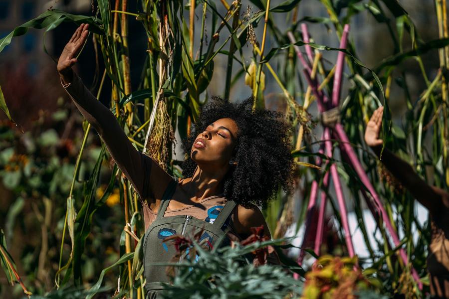 A Black woman, with natural hair and wearing olive overalls, holds her hand up to a spotlight. Another hand is raised next to her.