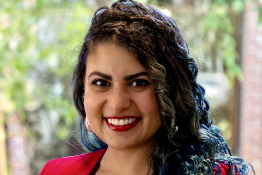 Outside, a woman with long curly, brown hair, smiles. She wears a red blazer that matches her lipstick and a paisely blouse.