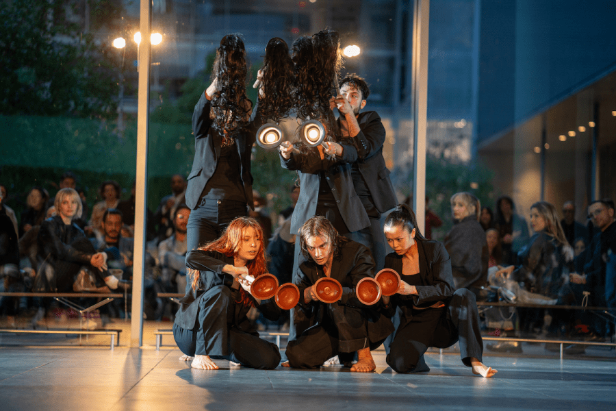 Three performers stand holding dark wigs on sticks. Crouching in front of them three performers hold toilet plungers.