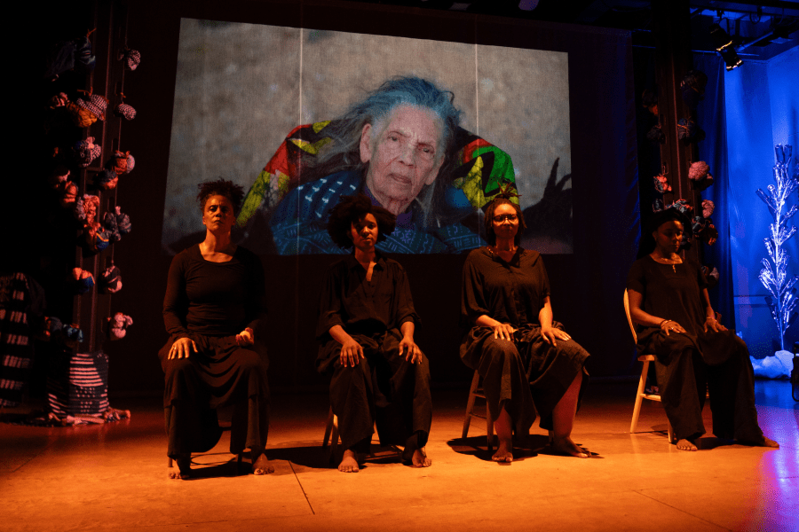 Four performers dressed in black sit on a stage. Behind them is a projection of an elderly person.