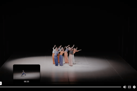 A group of dancers huddle in a spotlight, in a much larger stage.