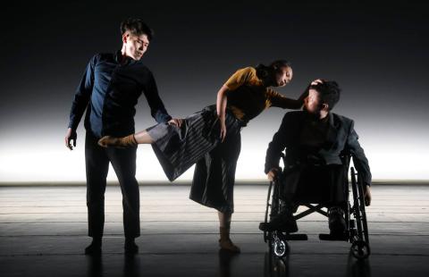 A male dancer holds the leg of a female dancer, who leans over a male dancer in a wheelchair.