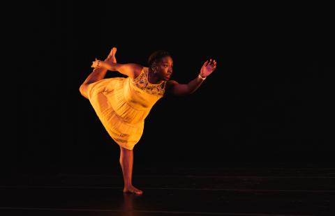 A Black woman holds her leg up and stands on one. She leans forward and holds her other arm out. She's in a yellow dress on a dark stage.