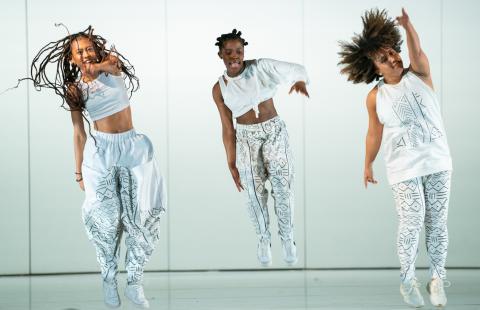 Three Black women jump and two point in different directions. They wear silver and are in front of a grey background with studio lighting.