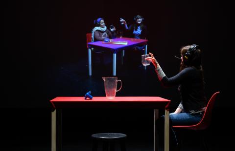 A person at a table holding a glass looking at a projection of  two people at another table holding glasses.