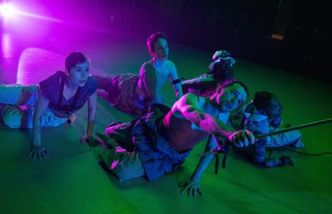 Laying on a stage, in green, purple, and blue light, five dancers crawl. One holds a pole that goes out of the frame.