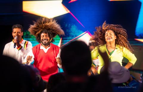 On a stage, and before a silhoutted audience, three folks, wearing bright colors, move their bodies and hit drums. A woman's hair is caught mid motion.