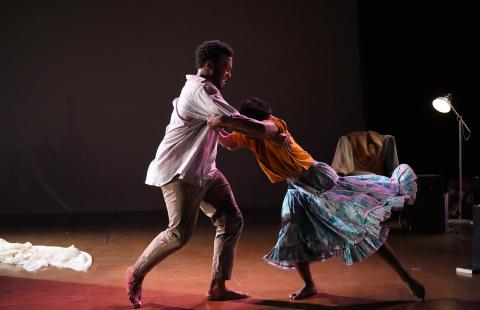 On a stage, with a chair and lamp, two Black folks dance. One of the dancer's dress spins out and away from her.