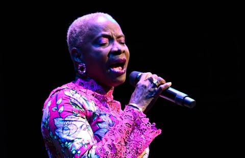 A Black woman in a pink and green dress. She sings into a microphone under a spotlight.
